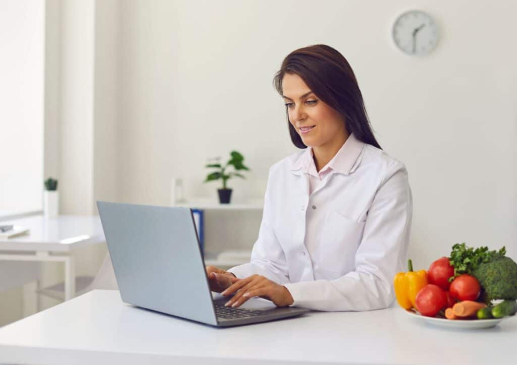What Is A Nutrition Coach? 1 A woman in a white lab coat sits at a table, focused on her laptop, in a professional setting.