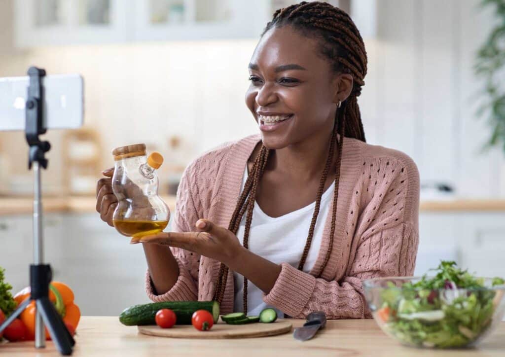 What Is A Nutrition Coach? 2 A woman holds a jar of olive oil in one hand and a camera in the other, smiling at the camera.