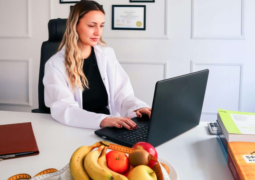 What Is A Nutrition Coach? 6 A woman in a white lab coat sits at a desk, focused on her laptop, in a professional office setting.