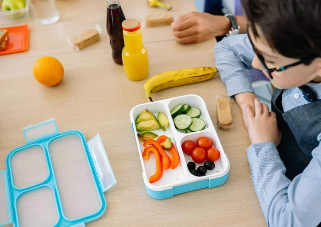 What Is School Wellbeing? 2 A child sitting at a table, opening a colorful lunch box filled with food items.