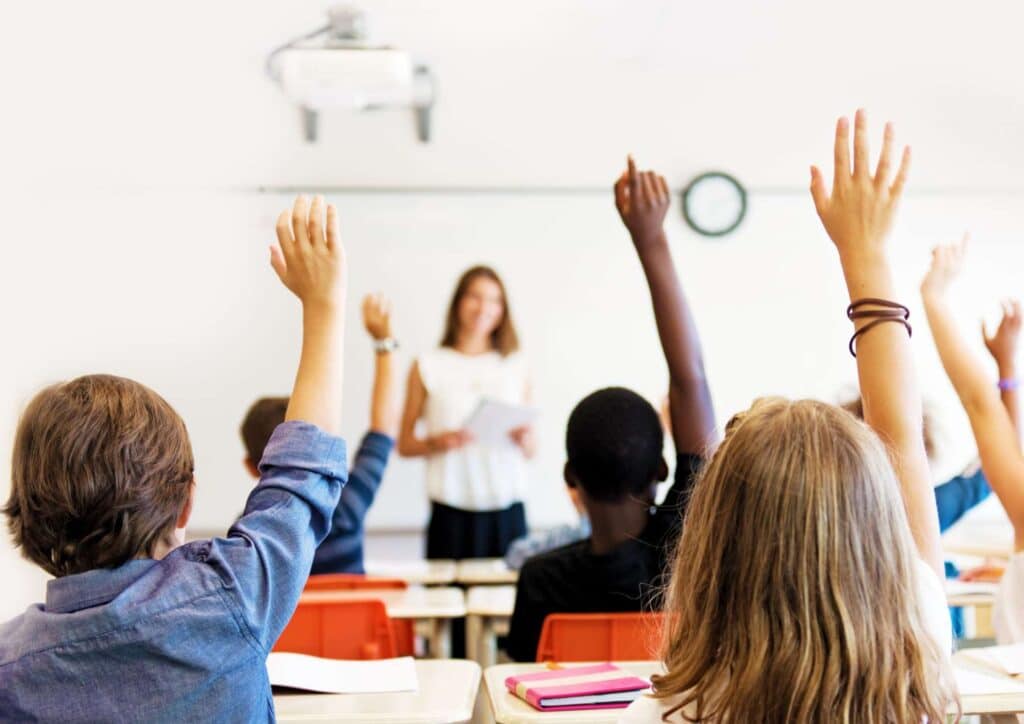 What Is School Wellbeing? 3 A group of children in a classroom eagerly raising their hands to answer a question from the teacher.