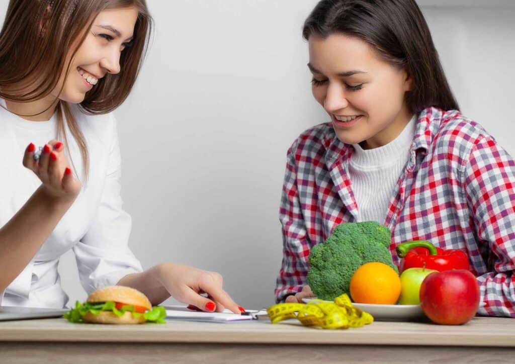 What Is A Weight Loss Nutritionist? 5 Two women seated at a table, engaged in conversation with a laptop and a plate of food in front of them.