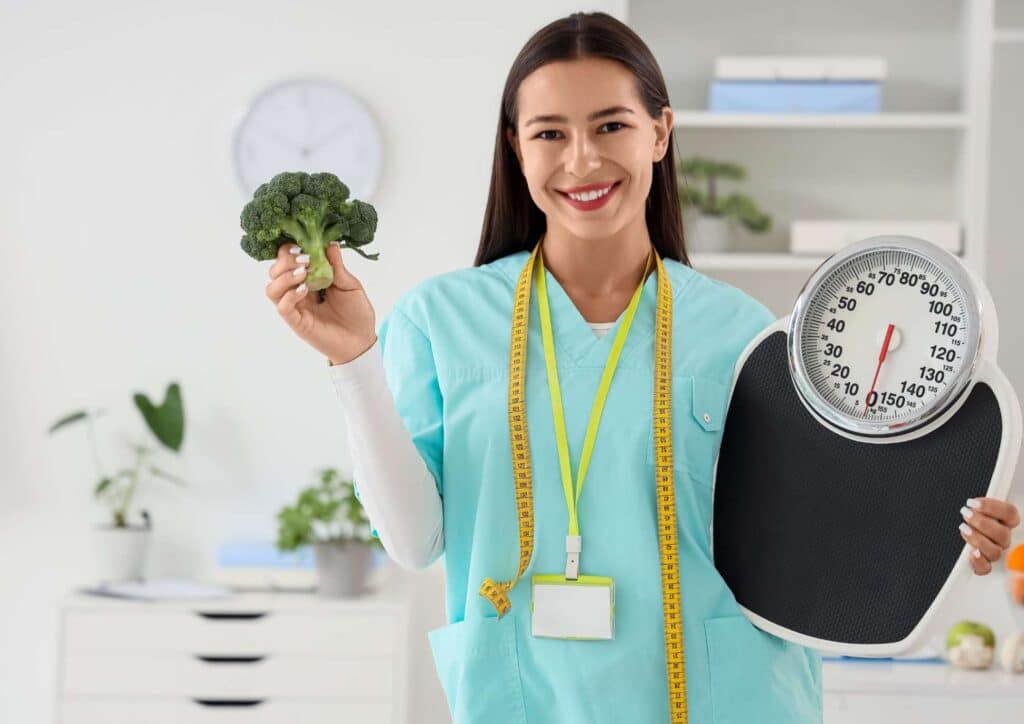 What Is A Weight Loss Nutritionist? 4 A woman in scrubs holds a scale and a broccoli, symbolising health and nutrition in a medical or wellness setting.
