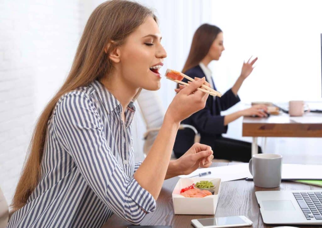 Corporate Nutrition Speaker Workplace Benefits 8 A woman sitting at a desk enjoys sushi, with a focused expression and a plate of sushi in front of her.