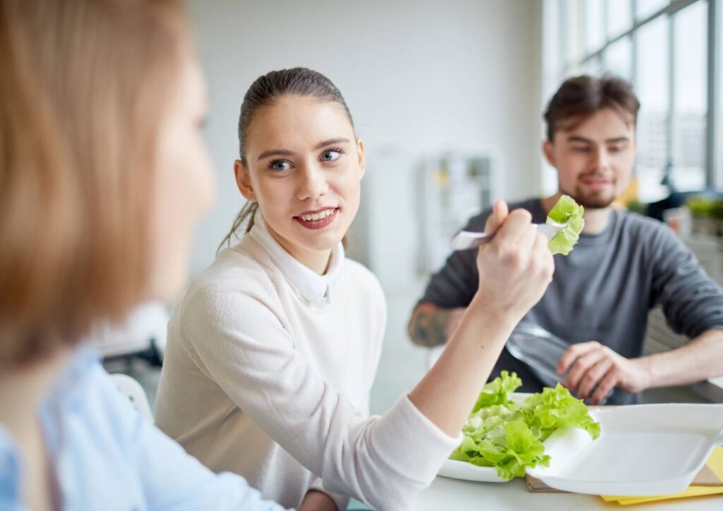 Corporate Nutrition Speaker Workplace Benefits 6 A woman eats lettuce while two men observe her, engaged in conversation.