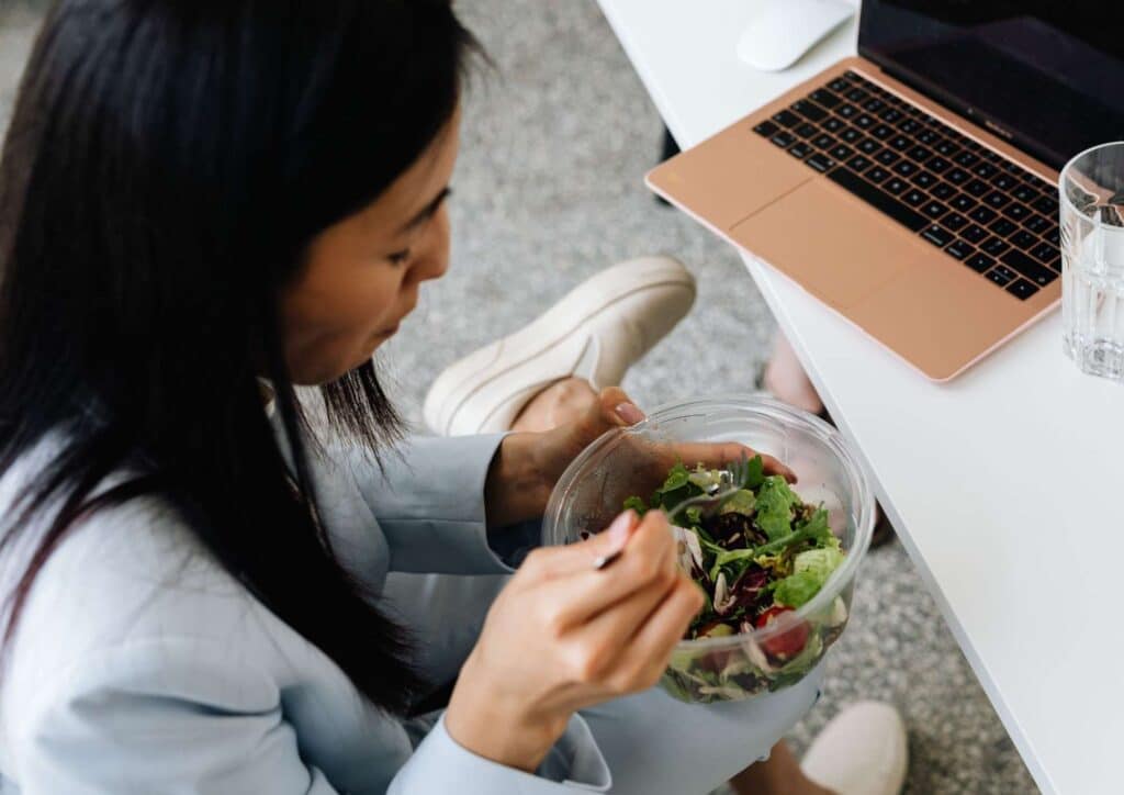 Corporate Nutrition Speaker Workplace Benefits 3 A woman enjoying a salad while working on her laptop at a desk, focused on her meal and screen.