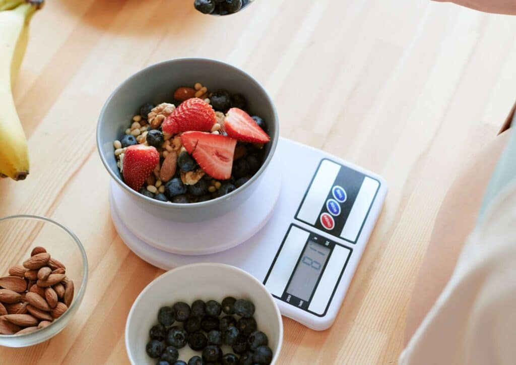 What Is The Perfect Diet? 4 A person measures a bowl filled with assorted fruits and nuts on a digital scale in a kitchen setting.