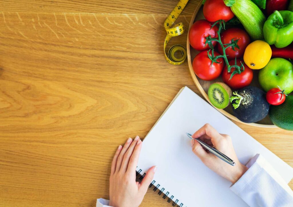 What Does a Nutritionist Do? 3 A woman writes in a notebook beside a bowl filled with fresh vegetables, indicating a focus on healthy cooking or meal planning.
