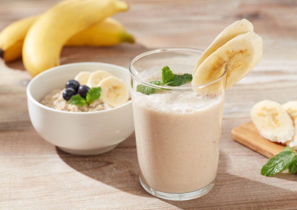 High Protein Banana Smoothie For Weight Loss 7 A banana smoothie with oats sits in a glass on a wooden table, surrounded by fresh bananas and a rustic setting.