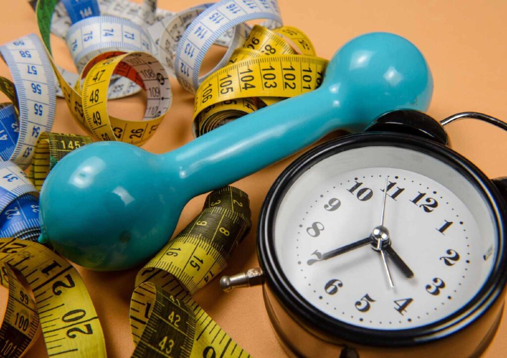 What Is A Sports Nutritionist? 2 A clock, measuring tape, and a blue dumbbell arranged on a flat surface, symbolising fitness and time management.