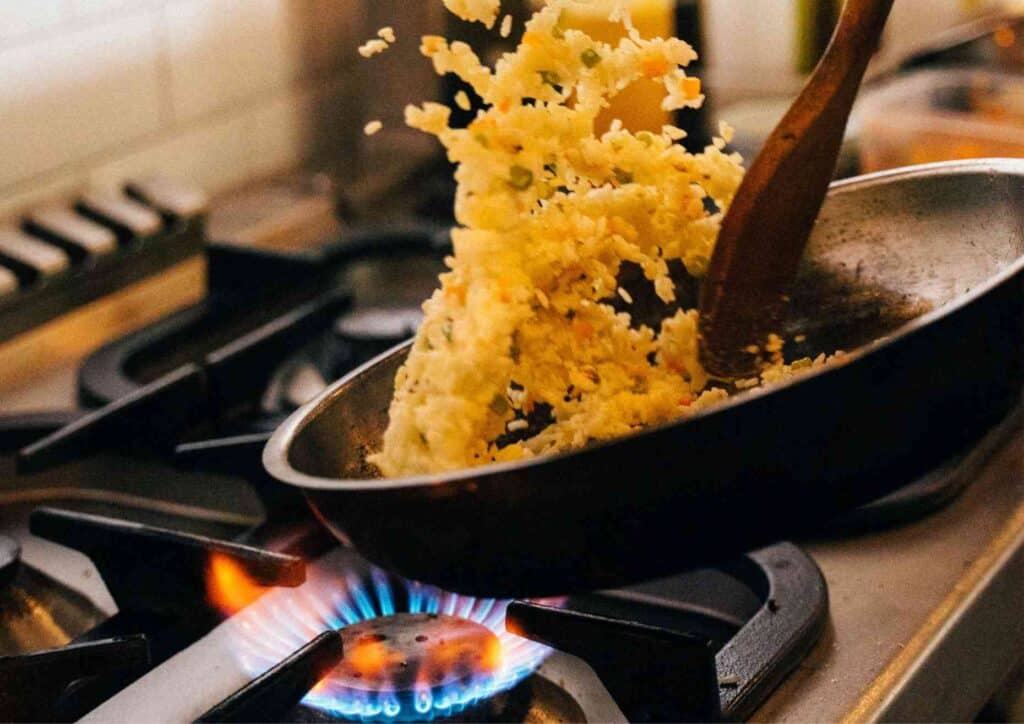 Is Rice Good for Weight Loss? A Nutritionist Explains 4 A frying pan on a gas stove with a blue flame visible underneath, tossing rice or noodles mid‑air with a wooden spatula during cooking.