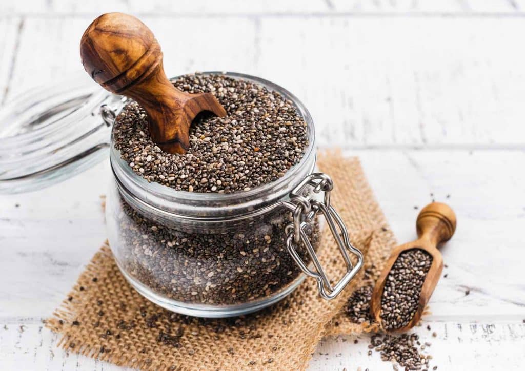 Do Chia Seeds Help With Weight Loss? 7 Chia seeds in a glass jar beside a wooden spoon on a light surface.