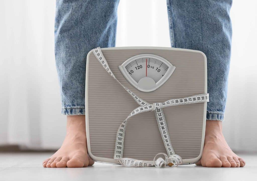 How to Break a Weight Loss Plateau, a Nutritionist's Approach 21 A person stands on a scale, holding a tape measure, indicating a focus on weight and body measurements.