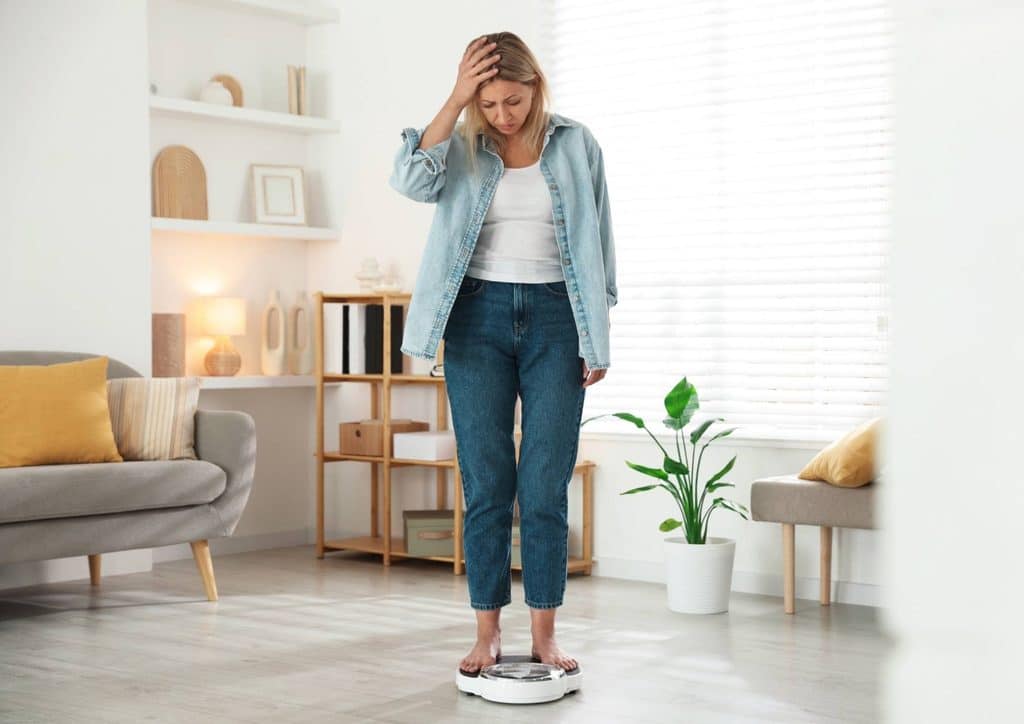How to Break a Weight Loss Plateau, a Nutritionist's Approach 17 A woman stands on a scale in a well-lit room, looking down at the display with a focused expression.