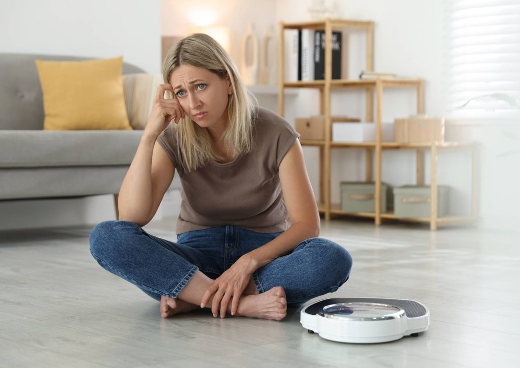How to Break a Weight Loss Plateau, a Nutritionist's Approach 12 A woman sits on the floor, observing a robotic vacuum cleaner as it navigates around her living space.