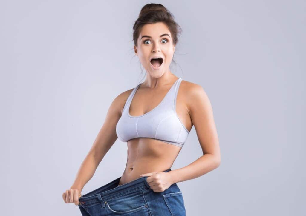 Does Lemon Water Help With Weight Loss? 2 A woman in a white top and blue jeans holds up her jeans, smiling confidently at the camera.