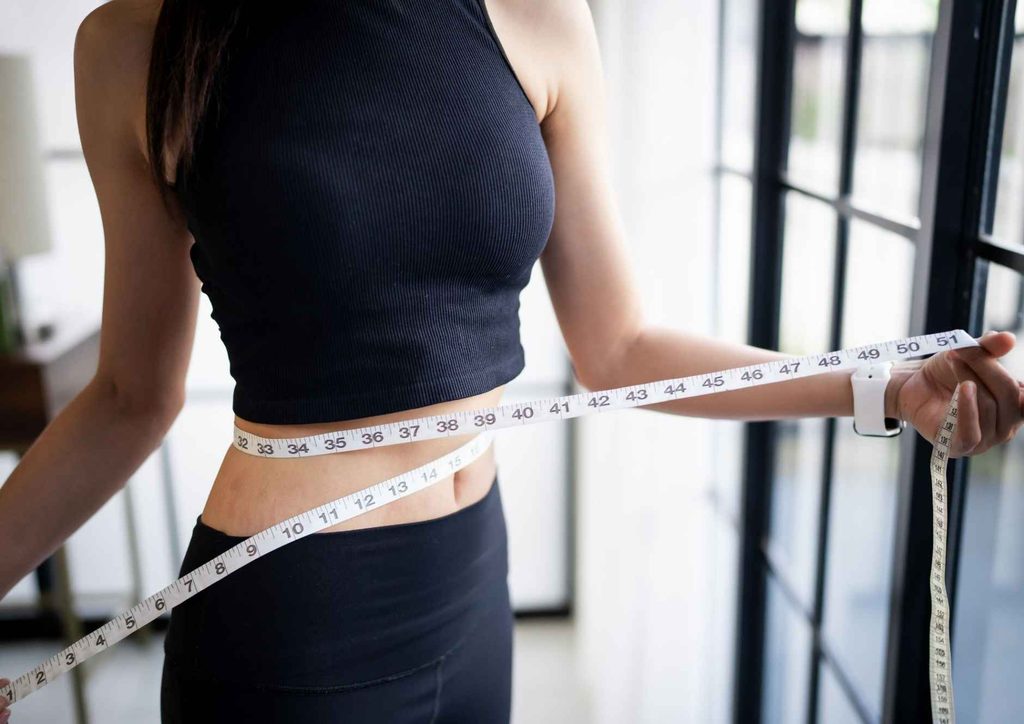 Are Oats Good for Weight Loss? 4 A woman measuring her waist with a tape measure, focusing on her midsection in a well-lit room.