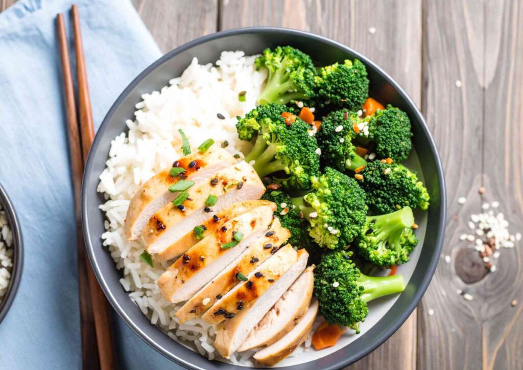 Is Rice Good for Weight Loss? A Nutritionist Explains 3 A bowl containing sliced grilled chicken breast served over white rice with broccoli on the side, placed on a wooden surface with chopsticks nearby.