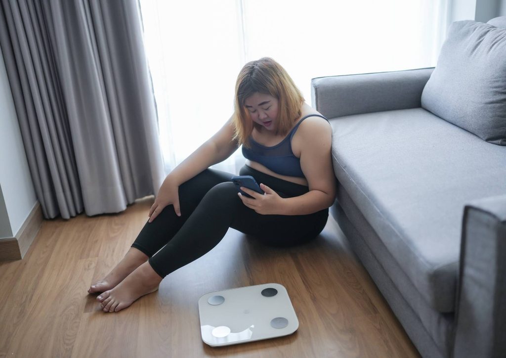 How to Break a Weight Loss Plateau, a Nutritionist's Approach 8 A woman sits on the floor beside a scale, looking contemplative and focused on her weight measurement.