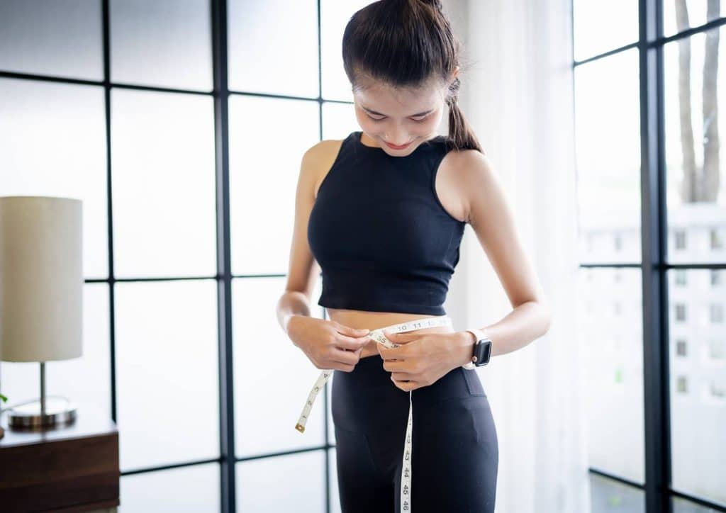 Do Saunas Help With Weight Loss? What Actually Happens 17 Asian woman measuring her waistline with a tape measure in a bedroom setting, reflecting personal health and fitness.