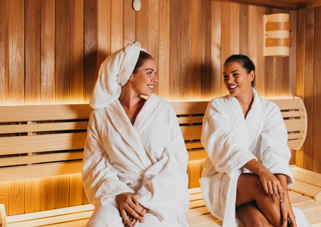 Do Saunas Help With Weight Loss? What Actually Happens 16 Two women in white robes relax on a wooden bench inside a sauna, surrounded by warm, steamy air.
