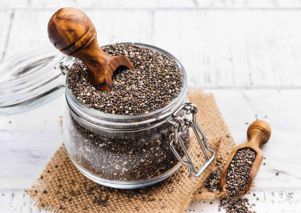 Do Chia Seeds Help With Weight Loss? 18 Chia seeds in a glass jar with a wooden spoon resting on the rim, showcasing their small, black, and white textures.