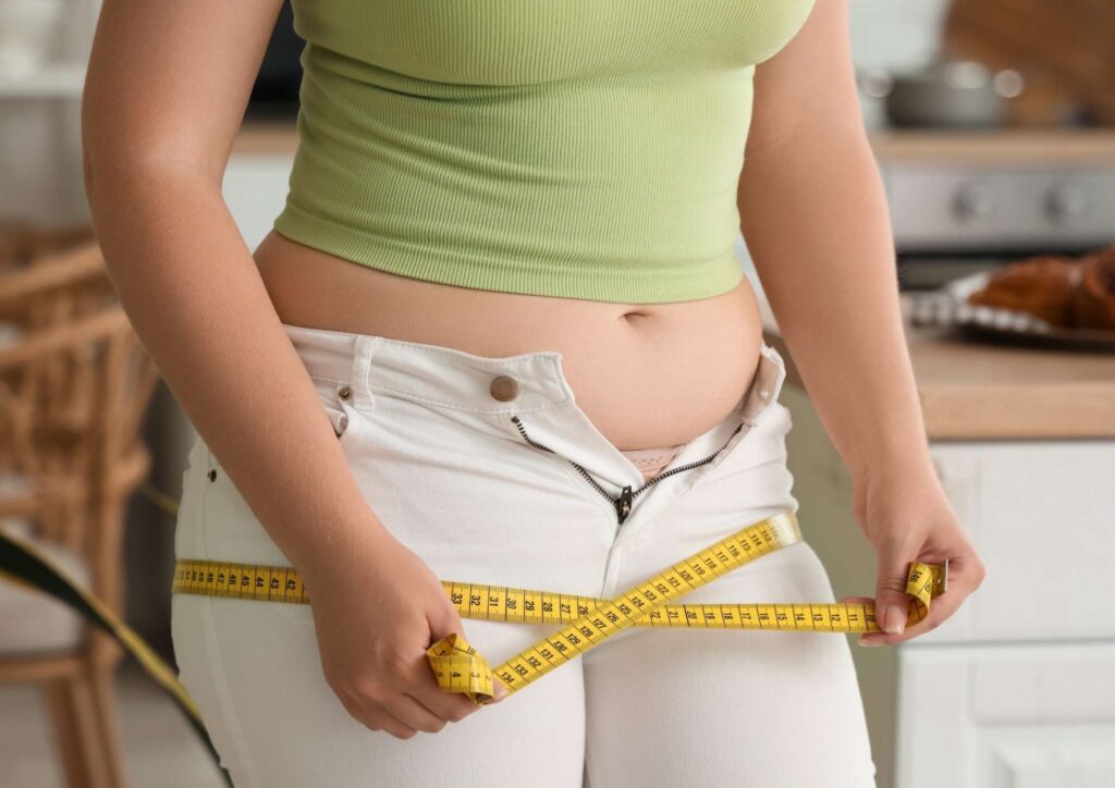 How to Break a Weight Loss Plateau, a Nutritionist's Approach 27 A woman in a green top measures her waist with a tape measure, focusing on her midsection.