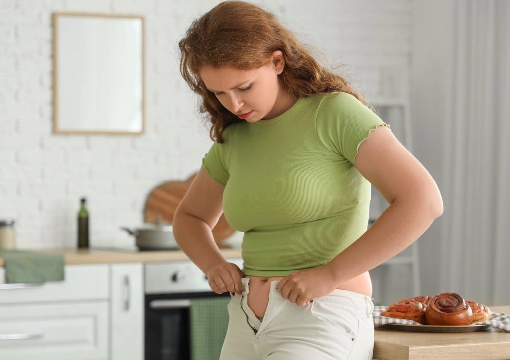 How to Break a Weight Loss Plateau, a Nutritionist's Approach 26 A woman in a green shirt is standing and holding her stomach, appearing to be in discomfort or pain.