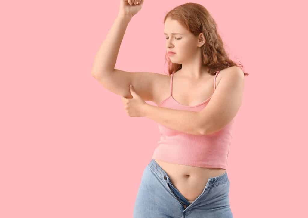 How to Break a Weight Loss Plateau, a Nutritionist's Approach 16 A woman in a pink top and jeans proudly displays her toned arms, smiling at the camera.