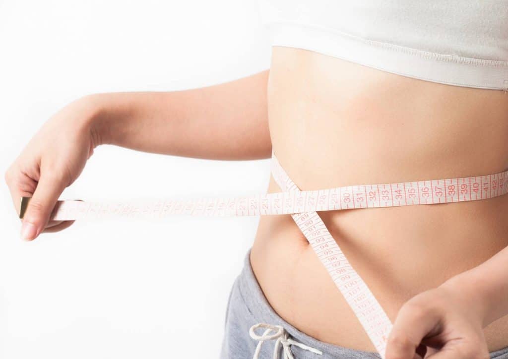 How to Break a Weight Loss Plateau, a Nutritionist's Approach 22 A woman measures her waist with a tape measure, focusing on her midsection in a well-lit room.