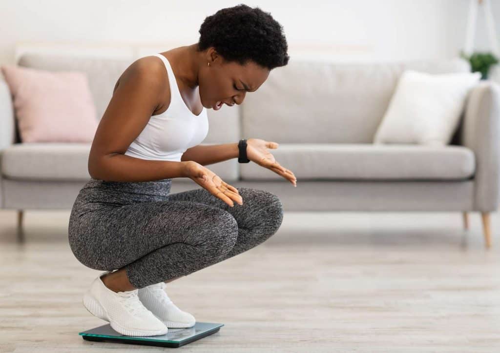 How to Break a Weight Loss Plateau, a Nutritionist's Approach 14 A woman squats on a scale, checking her weight while maintaining a focused expression.