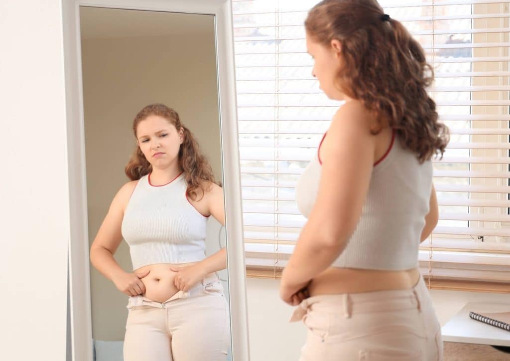 How to Break a Weight Loss Plateau, a Nutritionist's Approach 11 A woman stands in front of a mirror, gazing at her reflection with a thoughtful expression.