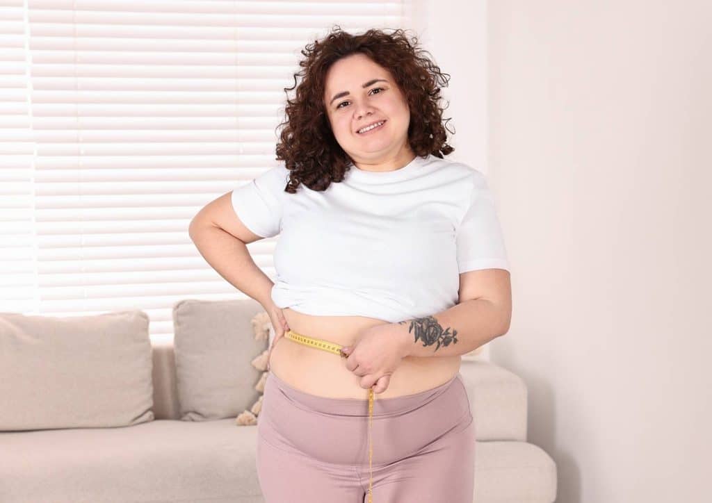 Do Saunas Help With Weight Loss? What Actually Happens 23 A woman stands in front of a couch, hands resting on her stomach, appearing contemplative and relaxed.