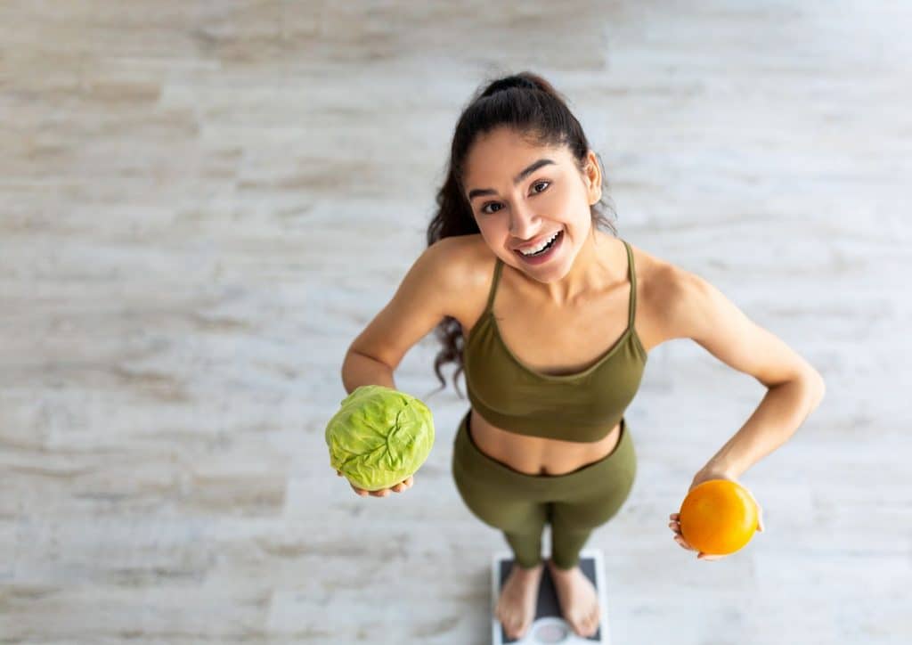 The Best Foods for Weight Loss 1 A woman in green sports attire holds an orange in one hand and a green apple in the other, smiling confidently.