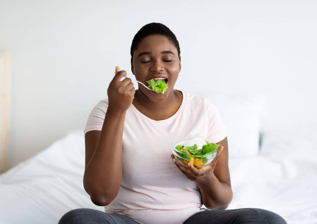The Best Foods for Weight Loss 13 A woman sitting on a bed, enjoying a bowl of salad, with a relaxed expression on her face.