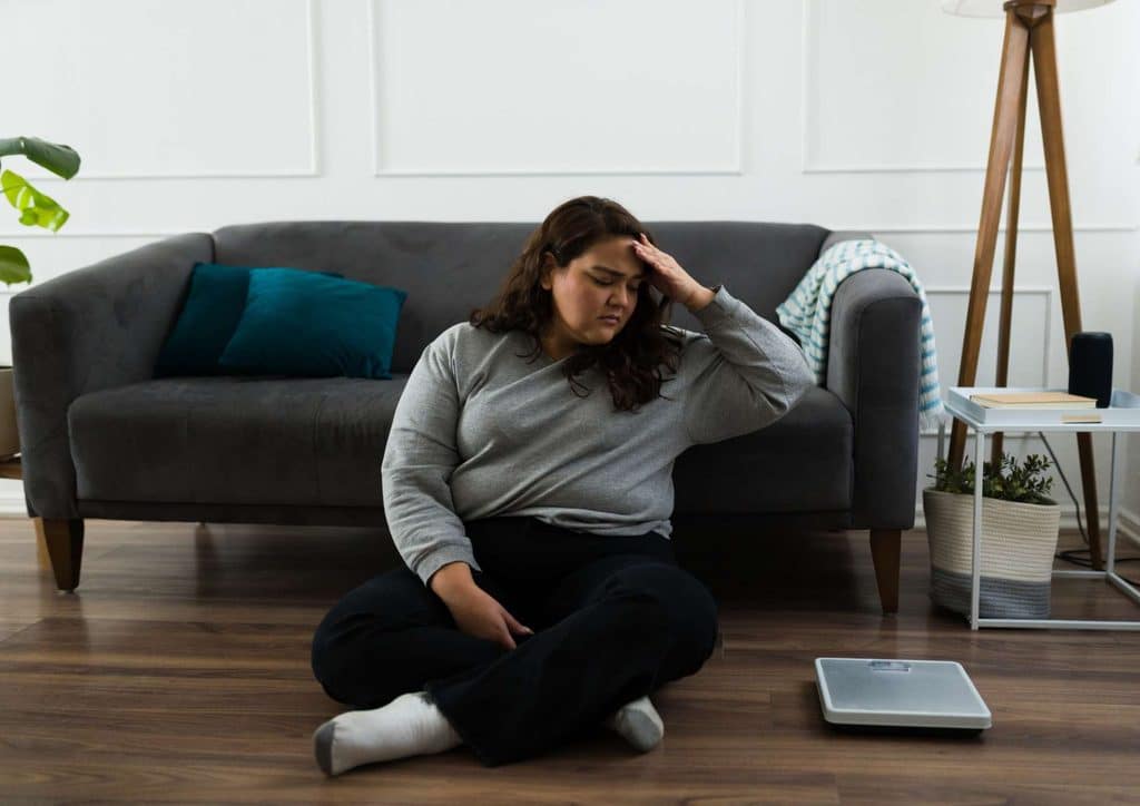 How to Lose Belly Fat: What a Nutritionist Recommends 8 A woman sits on the floor, her head in her hands, conveying a sense of distress or deep contemplation.