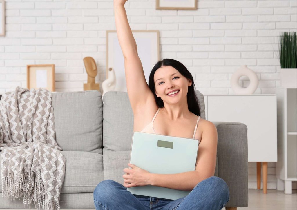 How to Lose Belly Fat: What a Nutritionist Recommends 19 A woman sitting on a couch, smiling while holding a weight scale in her hands.