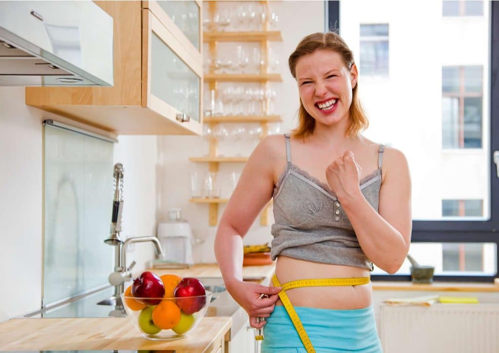 How to Lose Belly Fat: What a Nutritionist Recommends 17 A woman measures her waist with a tape measure while standing in a kitchen, surrounded by cooking utensils.