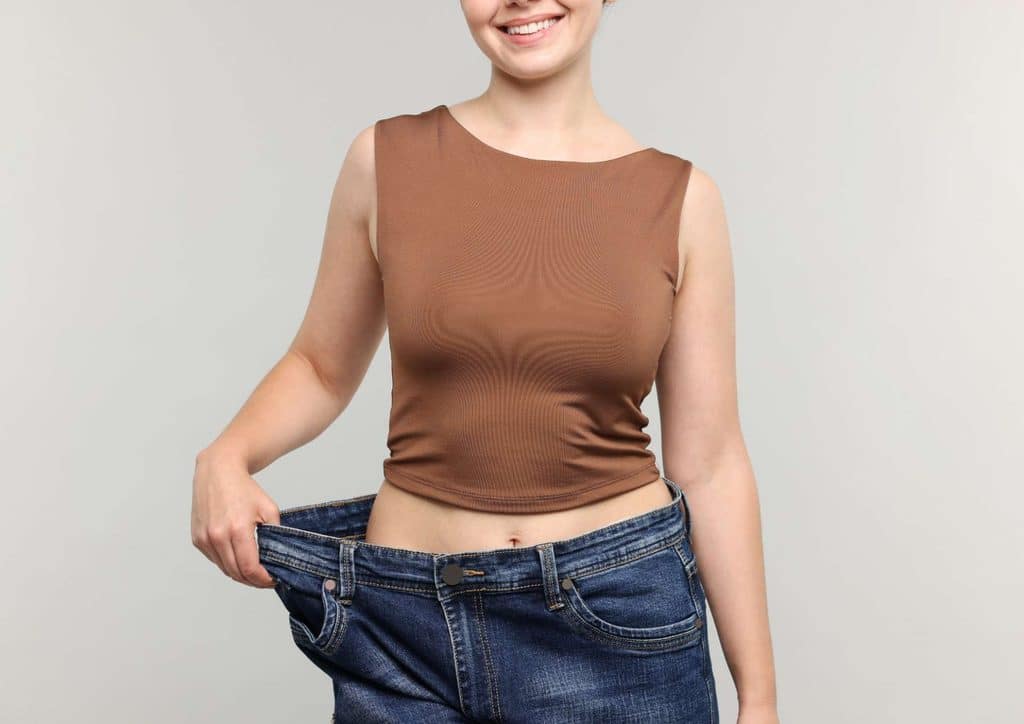 How to Lose Belly Fat: What a Nutritionist Recommends 28 A woman in a brown top and jeans is standing, holding her stomach with a concerned expression.