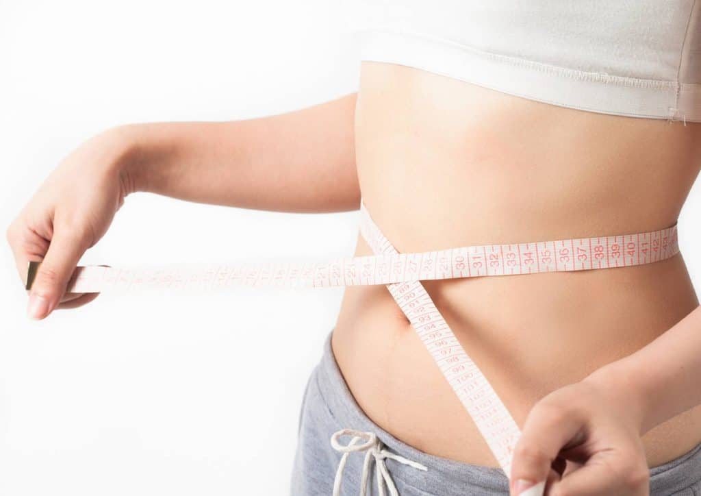 How to Lose Belly Fat: What a Nutritionist Recommends 22 A woman measures her waist with a tape measure, focusing on her midsection in a well-lit room.