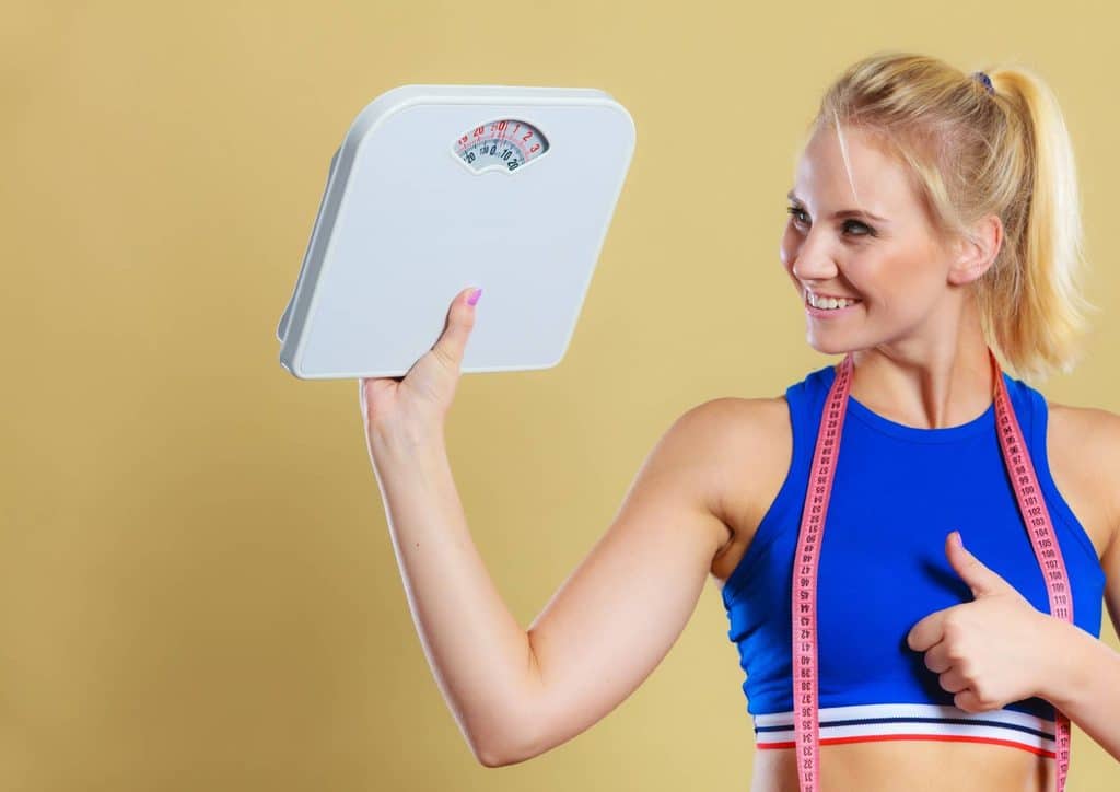 How to Lose Belly Fat: What a Nutritionist Recommends 14 A woman in a sports bra and shorts stands holding a weight scale, looking focused and determined.