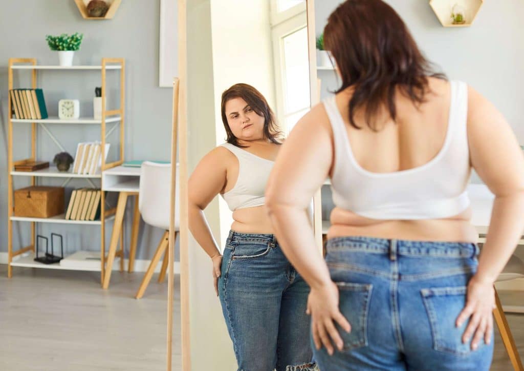 How to Lose Belly Fat: What a Nutritionist Recommends 27 A woman in a white shirt and blue jeans gazes at her reflection in a mirror.