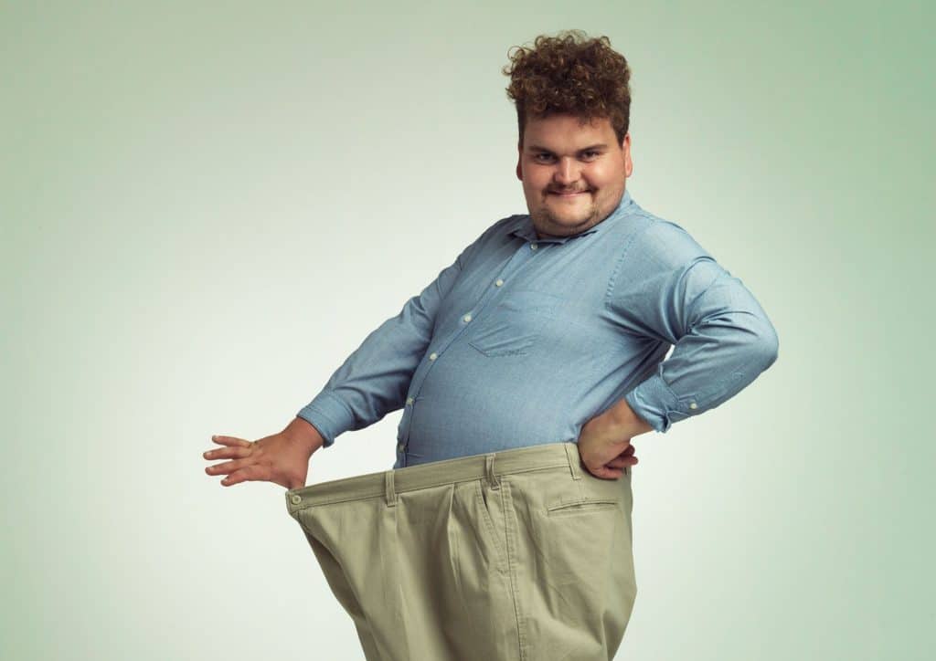 The Best Foods for Weight Loss 25 A man with a large belly is holding up his pants, standing casually with a neutral expression.