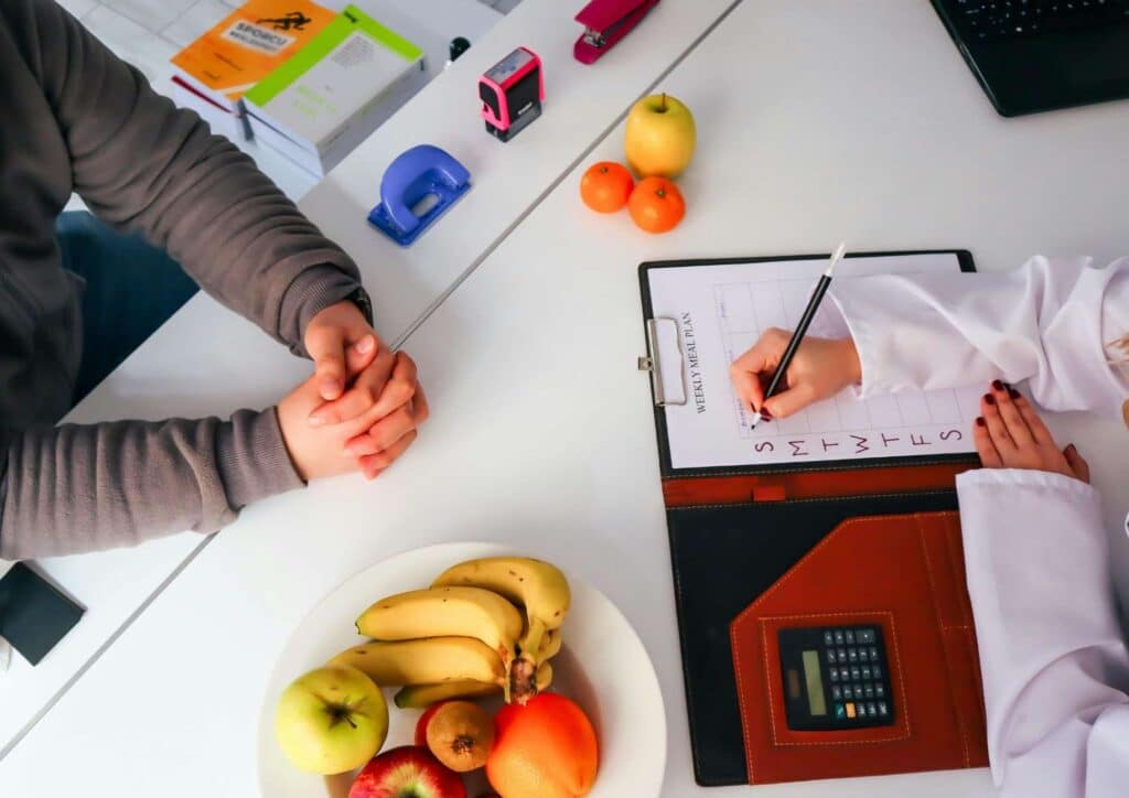 Nutritionist vs Clinical Nutritionist: What's the Difference? 4 A nutritionist writes on a clipboard while a patient sits at a table with fruit and a notebook, engaged in a consultation.