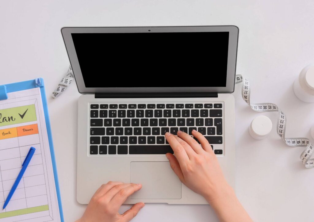 Nutritionist vs Clinical Nutritionist: What's the Difference? 1 A woman's hands typing on a laptop, with a notebook and a bottle of pills nearby on a wooden surface.