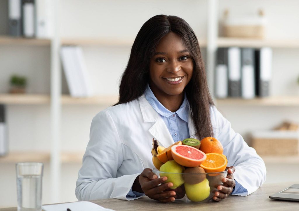 Clinical Nutritionist vs Nutritionist: What's the Difference 16 A woman in a lab coat smiles while holding a bowl filled with various fresh fruits.