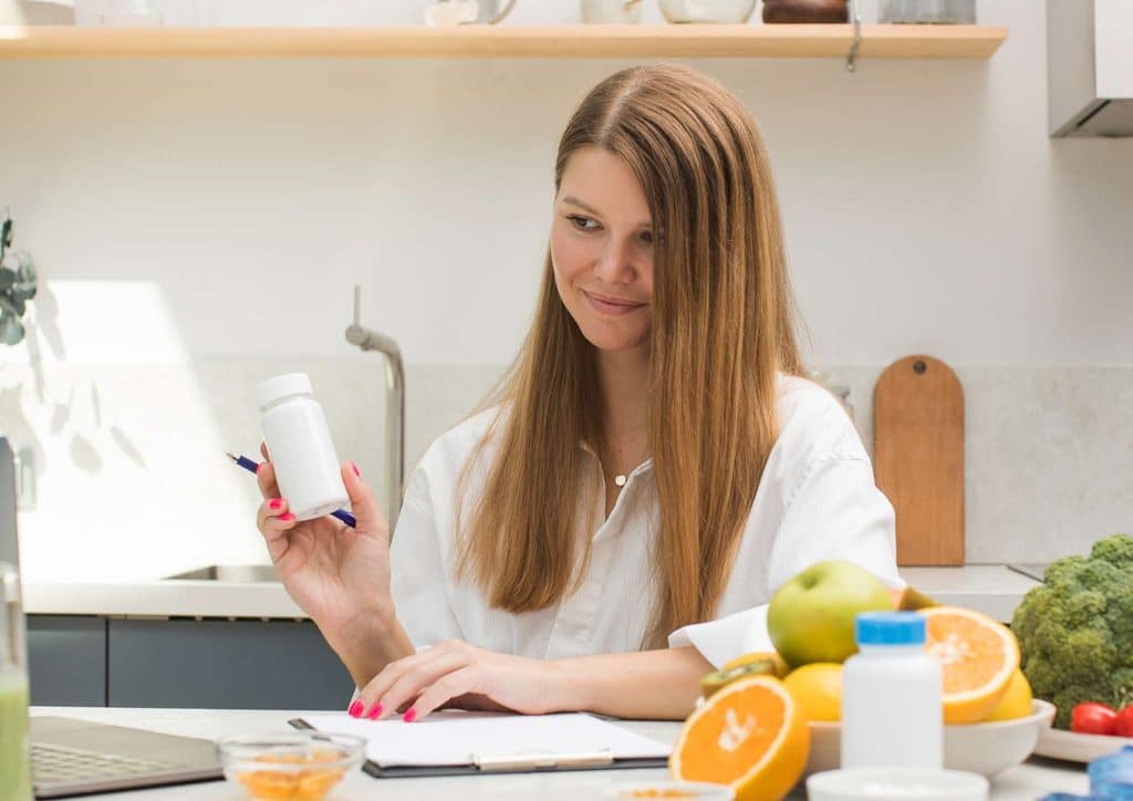 Clinical Nutritionist vs Nutritionist: What's the Difference 9 A woman sits at a table with a bottle of vitamins and an assortment of fresh fruit nearby.