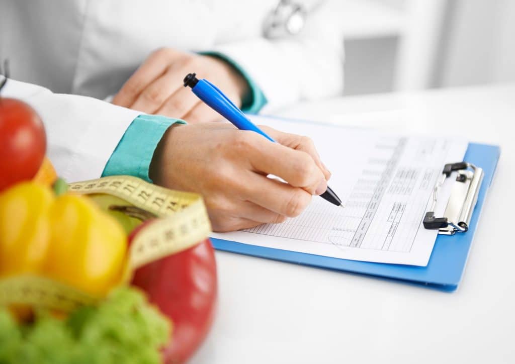 Clinical Nutritionist vs Nutritionist: What's the Difference 1 A doctor writes on a clipboard while surrounded by fresh fruits and vegetables, emphasising healthy eating