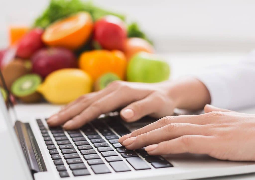 Clinical Nutritionist vs Nutritionist: What's the Difference 6 A person typing on a laptop, surrounded by various fruits and vegetables in the background.