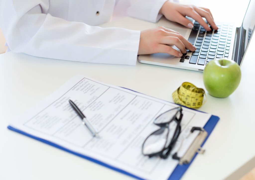 Nutritionist vs Clinical Nutritionist: What's the Difference? 7 A person seated at a desk, working on a laptop, with a red apple placed beside the computer